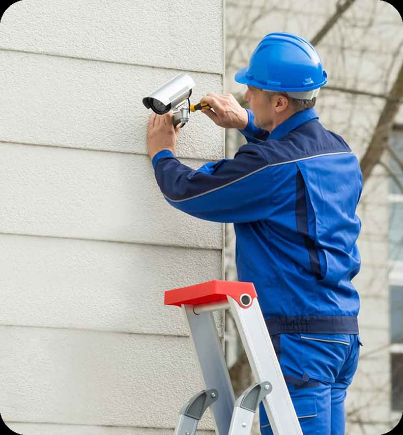 A man in blue overalls repairs a camera mounted on a wall,